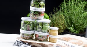 Three jars of fresh layered salad and a small jar of dressing, decoratively arranged on a wooden board with fresh herbs in the background.