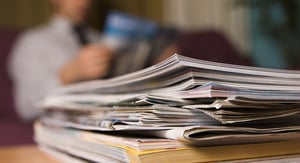 Ein Stapel Zeitschriften liegt auf einem Tisch im Vordergrund, während im unscharfen Hintergrund eine Person sitzt und ein Magazin liest.