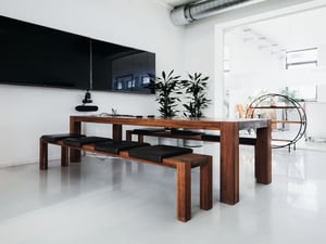 Modern eingerichteter Besprechungsraum mit einem großen Holztisch, passenden Bänken mit schwarzen Sitzpolstern, einem Flachbildschirm an der Wand und Pflanzen im Hintergrund.